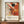 May include: Framed Jackson Pollock exhibition poster featuring abstract expressionist artwork with dynamic orange, red, black and yellow paint splatters and brushstrokes on cream background. Exhibition details read January 10-20, 1956, 8th Avenue, New York City, "A Journey Through Chaos." Displayed on wooden sideboard beside white ceramic vase and stacked books against cream wainscoted wall.