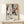 May include: Framed Jackson Pollock exhibition poster with abstract black paint splatters and red accents on cream background, displaying in light wood frame on white wooden console table beside small white gift box with twine and greenery against pale wall