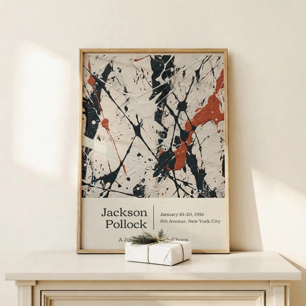 May include: Framed Jackson Pollock exhibition poster with abstract black paint splatters and red accents on cream background, displaying in light wood frame on white wooden console table beside small white gift box with twine and greenery against pale wall