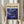 May include: Person holding framed abstract art print with swirling deep blue and purple cosmic patterns, scattered gold and white dots resembling stars or paint splatters. Cream mat border, light wood frame. Dried pampas grass stems visible on left side. Soft beige clothing and neutral curtained background create minimalist styling.