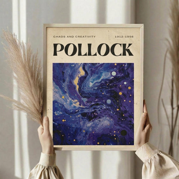 May include: Person holding framed abstract art print with swirling deep blue and purple cosmic patterns, scattered gold and white dots resembling stars or paint splatters. Cream mat border, light wood frame. Dried pampas grass stems visible on left side. Soft beige clothing and neutral curtained background create minimalist styling.
