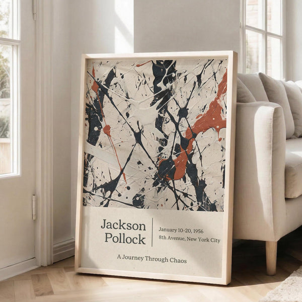 May include: Abstract expressionist artwork poster with black paint splatters, drips and orange-red accents on cream background, displayed in light wood frame leaning against white wall next to beige upholstered chair in bright, naturally-lit room with large windows and hardwood floors.