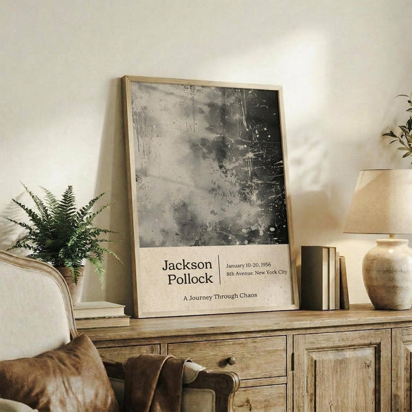 May include: Abstract black and gray splattered artwork in light wood frame, displayed on rustic wooden sideboard with fern plant, ceramic vase, and books. Neutral living room setting with beige chair and soft natural lighting. Exhibition-style poster features textured paint splatters and drips in monochromatic tones.