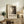 May include: Framed vintage-style exhibition poster with abstract black and white splattered artwork displayed on rustic wooden console table beside fern plant, cream armchair, ceramic vase, and stacked books in neutral-toned living room setting.