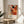 May include: Abstract expressionist art poster in light wood frame displaying vibrant orange, red, and black paint splatters on cream background with exhibition text below. Positioned on round wooden side table next to beige upholstered chair, with plant leaves visible and natural sunlight casting shadows on white wall behind.