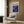May include: Abstract swirling blue and purple poster with cosmic design featuring scattered dots and flowing paint-like patterns, displayed in light wood frame on wood paneled wall above beige upholstered armchair with geometric patterned pillow, alongside brass floor lamp and dark round side table with open book.