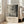 May include: A framed poster displaying abstract black and gray paint splatters and drips on white background, with text reading "Jackson Pollock" and exhibition details below. The cream-colored wooden frame leans against a white wall next to a beige upholstered sofa in a bright, minimalist living room with large windows and hardwood floors.
