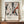 May include: Abstract art poster with black paint splatters and orange-red accents on cream background, displayed in thin brown frame on wooden sideboard. White ceramic vase and stacked books sit nearby. Exhibition-style text below artwork shows artist name and exhibition details. Wall has white wainscoting panels.