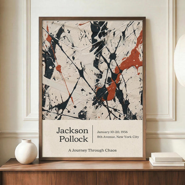 May include: Abstract art poster with black paint splatters and orange-red accents on cream background, displayed in thin brown frame on wooden sideboard. White ceramic vase and stacked books sit nearby. Exhibition-style text below artwork shows artist name and exhibition details. Wall has white wainscoting panels.