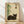 May include: Framed artwork displaying a sleeping brown tabby cat beside green bamboo stalks on cream background with Japanese characters and red stamp seal, shown in light wood frame leaning against white wall on wooden floor with soft natural lighting