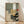 May include: Framed abstract geometric artwork featuring sage green and cream rectangular blocks with silhouette of black pine tree in foreground and orange circle accent, displayed against white textured wall on light wood floor with diagonal shadows