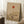 May include: Framed minimalist artwork leaning against white wall featuring orange-red sun, bare tree with delicate branches, and layered beige hills in Japanese-inspired style. Scene shows cozy bedroom setting with white coffee cup on wooden block, cream textured rug, and soft beige bedding with natural window light casting shadows.