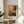May include: Abstract expressionist art poster with vibrant orange, red, blue and black paint splatters and drips on white background, displayed in thin wooden frame on round wooden side table next to beige upholstered chair, with plant leaves visible in soft natural lighting and neutral wall backdrop.