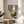May include: Framed abstract art print featuring geometric shapes in sage green, cream, mustard yellow, and gray with silhouette of windswept pine tree and small red circle accent, displayed on light wood round side table next to beige upholstered chair in bright room with plant shadows on white wall
