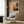 May include: Framed Jackson Pollock abstract art poster with vibrant orange, red, blue and black paint splatters displayed above a cream upholstered armchair with geometric pillow. Modern reading corner features brass floor lamp, dark side table with open book, and warm wood paneled wall. Natural lighting from window with sheer curtains illuminates the contemporary space.