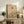 May include: Framed minimalist artwork displaying a red sun, curved line, bare tree silhouette, and beige hills on cream background, positioned on rustic wooden sideboard alongside fern plant, books, ceramic vase, and leather armchair in warm-toned living room setting.