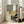 May include: Framed abstract artwork leaning against white wall next to beige upholstered chair in bright living room. Print features geometric blocks in sage green, cream, golden yellow and gray tones with silhouetted pine tree branches and coral red circle accent. Natural wood frame, warm sunlight streaming through windows onto hardwood floors.