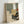 May include: Framed abstract art print featuring geometric shapes in muted green, beige, and gray tones with a black silhouette of a Japanese pine tree in the lower right corner and a small orange circle accent. The artwork is displayed in a light wood frame leaning against a white wall on a light surface, showing how it might look in a modern interior setting.