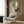 May include: Framed abstract artwork with geometric squares in sage green, warm beige, orange, and gray tones, featuring a black silhouette of a pine tree and red circle accent. Displayed above a cream upholstered armchair with geometric patterned pillow in a modern living room with wood paneled walls, brass floor lamp, and side table.