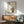 May include: Framed poster featuring a cute black cat with large yellow eyes and small red nose sitting next to bamboo stalks, with Japanese characters above. The artwork has a soft, watercolor-like style on cream background with brown wooden frame. Displayed above a light wood sideboard with white table lamp, next to a mint green mid-century modern armchair. Natural sunlight creates diagonal shadows across the white wall.
