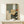 May include: Framed abstract artwork with geometric shapes in sage green, beige, and terracotta tones, featuring a silhouetted pine tree. The piece combines angular blocks of color with organic tree forms and includes a red circular sun element. Displayed on a light wood console table beside a small wrapped gift with natural twine and pine sprig against cream walls.