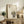 May include: Framed poster featuring a cute black cat with large yellow eyes and pink tongue sitting among bamboo stalks, with Japanese characters at top. Light wood frame displayed on rustic wooden sideboard alongside fern plant, books, ceramic vase, and brown leather furniture in warm, neutral-toned living room setting.