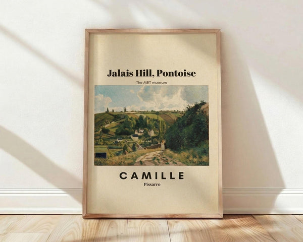 May include: Framed museum poster featuring Camille Pissarro's pastoral landscape painting showing rolling green hills, rustic buildings, and cloudy sky in soft earth tones. The cream-colored print displays "Jalais Hill, Pontoise" and "The MET museum" text above the artwork, with "CAMILLE Pissarro" below. Natural wood frame leans against white wall on light wooden floor with soft window shadows creating diagonal light patterns across the scene.