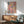 May include: Abstract expressionist artwork with vibrant paint splatters and drips in blue, pink, yellow, and red colors, displayed in wooden frame above mid-century modern credenza. Light green upholstered chair with wooden arms and small round coffee table visible in foreground. Natural sunlight creates geometric shadows on white wall, highlighting the energetic brushstrokes and layered paint textures of the colorful abstract composition.
