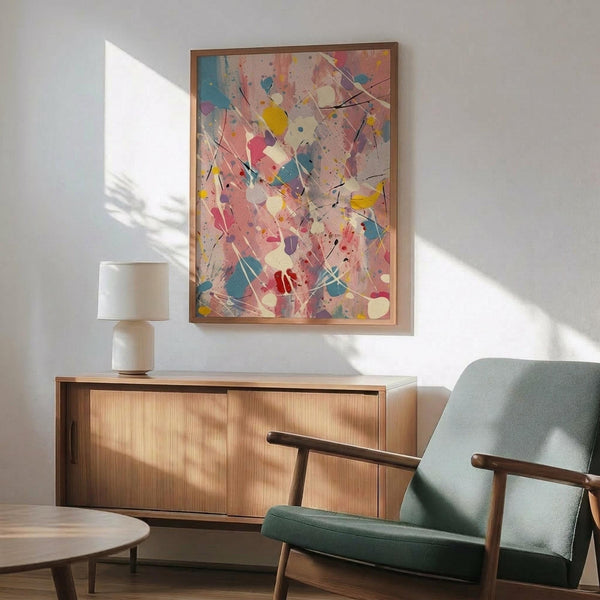 May include: Abstract expressionist artwork with vibrant paint splatters and drips in blue, pink, yellow, and red colors, displayed in wooden frame above mid-century modern credenza. Light green upholstered chair with wooden arms and small round coffee table visible in foreground. Natural sunlight creates geometric shadows on white wall, highlighting the energetic brushstrokes and layered paint textures of the colorful abstract composition.