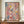 May include: Abstract expressionist artwork with vibrant paint splatters and drips in pink, blue, yellow, and white colors with black linear marks, displayed in thin brown frame on wooden sideboard next to white ceramic vase and stacked books in elegant room with cream walls and decorative molding.