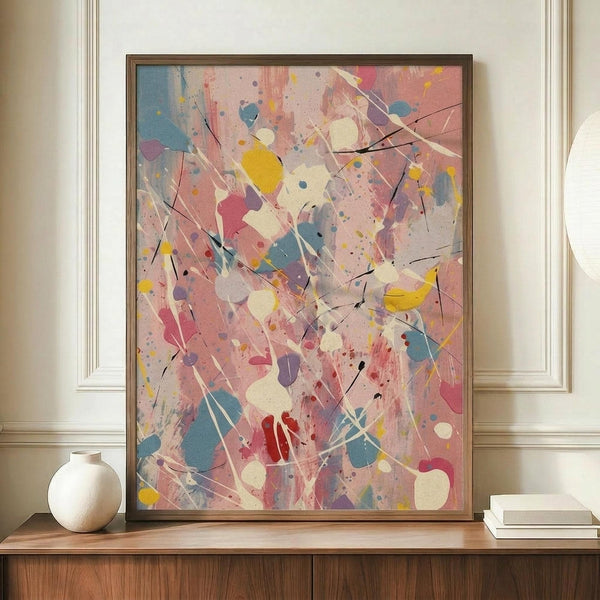 May include: Abstract expressionist artwork with vibrant paint splatters and drips in pink, blue, yellow, and white colors with black linear marks, displayed in thin brown frame on wooden sideboard next to white ceramic vase and stacked books in elegant room with cream walls and decorative molding.