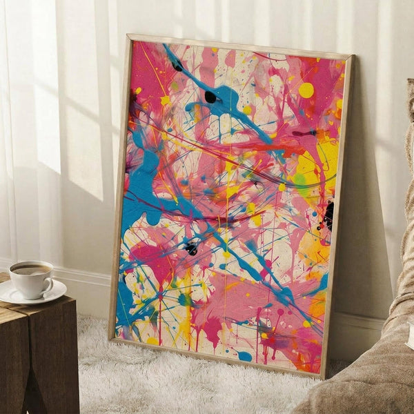 May include: Vibrant abstract expressionist artwork in light wood frame leaning against white wall. Features energetic paint splatters and drips in hot pink, turquoise blue, bright yellow, and black across cream background with dynamic brushstrokes and gestural marks. Displayed in cozy interior setting with white coffee cup on wooden block side table and textured cream throw blanket on white shag rug.