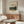 May include: Framed museum poster featuring Camille Pissarro's landscape painting of Jalais Hill, Pontoise displayed above a wood credenza in modern living room. Cream-colored poster shows rolling green countryside with buildings. Room has white curved chair, glass coffee table, brass floor lamp, and white table lamp. Minimalist Scandinavian-style interior with neutral tones and natural wood furniture.