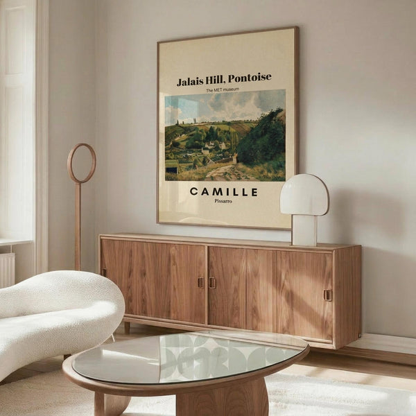 May include: Framed museum poster featuring Camille Pissarro's landscape painting of Jalais Hill, Pontoise displayed above a wood credenza in modern living room. Cream-colored poster shows rolling green countryside with buildings. Room has white curved chair, glass coffee table, brass floor lamp, and white table lamp. Minimalist Scandinavian-style interior with neutral tones and natural wood furniture.