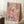 May include: Abstract expressionist artwork with vibrant paint splatters and drips in pink, blue, yellow, red, and white colors with black linear elements, displayed in a light wood frame leaning against white wainscoting beside a cozy armchair and small wooden side table with coffee cup on a soft cream rug
