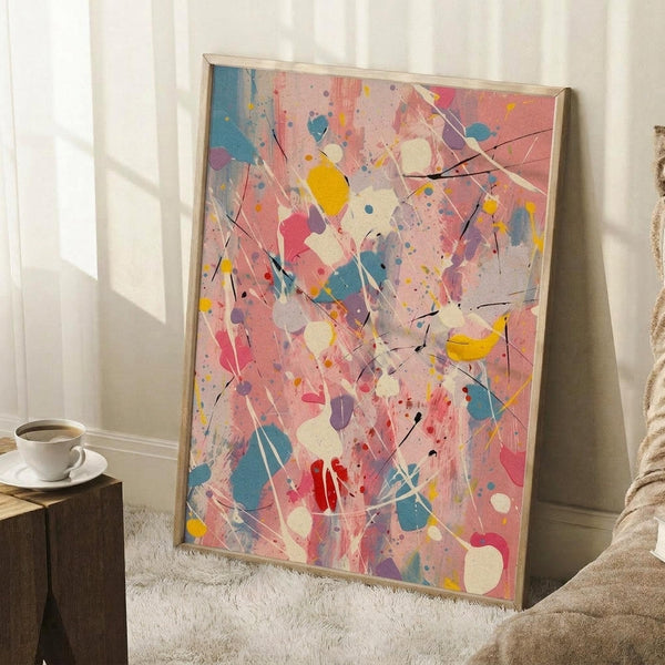 May include: Abstract expressionist artwork with vibrant paint splatters and drips in pink, blue, yellow, red, and white colors with black linear elements, displayed in a light wood frame leaning against white wainscoting beside a cozy armchair and small wooden side table with coffee cup on a soft cream rug