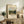 May include: Framed art print featuring a rural landscape with rolling hills, houses, and cloudy sky displayed on a wooden sideboard. The poster shows "CAMILLE" and "Pissarro" text below the pastoral scene. Styled with fern plant, books, ceramic vase, and neutral fabric chair in warm-toned room setting.