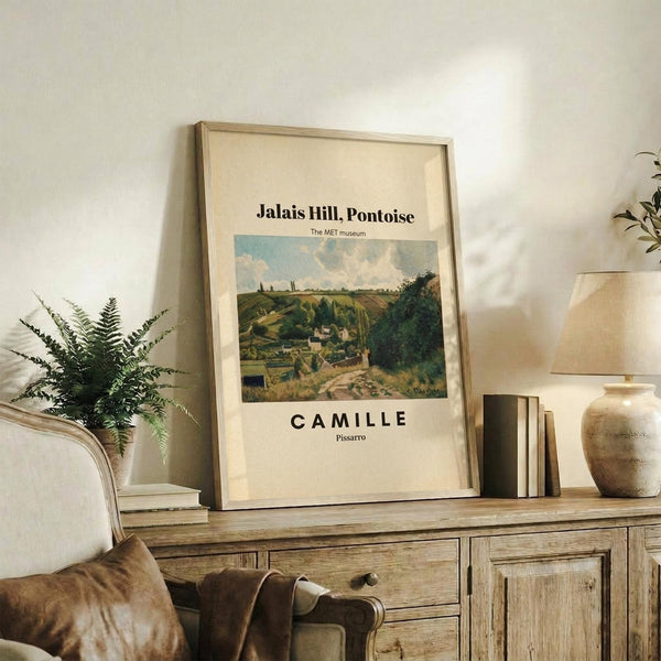 May include: Framed art print featuring a rural landscape with rolling hills, houses, and cloudy sky displayed on a wooden sideboard. The poster shows "CAMILLE" and "Pissarro" text below the pastoral scene. Styled with fern plant, books, ceramic vase, and neutral fabric chair in warm-toned room setting.