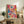May include: Abstract expressionist artwork with vibrant blue, pink, yellow and red paint splatters and dripped lines in light wood frame, displayed on rustic wooden sideboard alongside fern plant, ceramic vase, and books in neutral-toned living room setting with cream armchair visible.