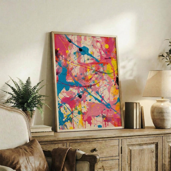 May include: Abstract expressionist artwork with vibrant blue, pink, yellow and red paint splatters and dripped lines in light wood frame, displayed on rustic wooden sideboard alongside fern plant, ceramic vase, and books in neutral-toned living room setting with cream armchair visible.