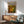 May include: Framed Jackson Pollock abstract art poster with vibrant orange, yellow, and green paint splatters and drips displayed above a mid-century modern wooden credenza with white ceramic lamp. Light blue upholstered armchair with wooden frame sits beside round coffee table in bright, naturally lit living room with white walls and warm wood flooring.