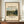 May include: Framed museum poster featuring Camille Pissarro's impressionist landscape painting of rolling green hills, village buildings, and cloudy sky in earth tones. Brown wooden frame displays on walnut sideboard beside white ceramic vase and stacked books against cream wainscoted wall.