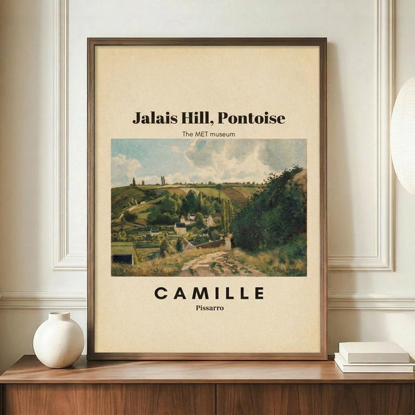 May include: Framed museum poster featuring Camille Pissarro's impressionist landscape painting of rolling green hills, village buildings, and cloudy sky in earth tones. Brown wooden frame displays on walnut sideboard beside white ceramic vase and stacked books against cream wainscoted wall.