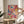 May include: Hands positioning a framed abstract splatter painting with vibrant pink, blue, yellow and red paint drips and splatters on cream background. Artwork sits on round wooden side table next to beige upholstered chair and potted plant in bright, naturally-lit room with white walls and curtains.