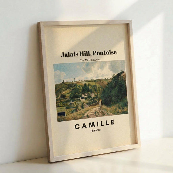 May include: Framed art print leaning against white wall showing landscape painting of rolling green hills with rural village, winding dirt road, and cloudy blue sky. Text reads "Jalais Hill, Pontoise" at top, "The MET museum" below, and "CAMILLE Pissarro" at bottom on cream background. Natural wood frame casts soft shadow on white surface.