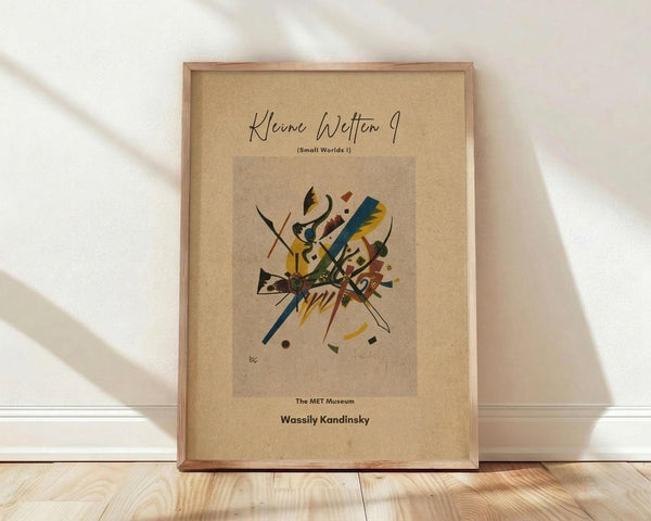 May include: Framed abstract art print with geometric shapes and bold colors including yellow, blue, red, and green on beige background, displayed in natural wood frame against white wall on light wooden floor with soft natural lighting creating diagonal shadows.