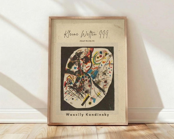 May include: Framed abstract art print in wooden frame leaning against white wall on light wood floor. Artwork features geometric shapes in vibrant colors - reds, blues, yellows, and greens with black lines and organic forms on cream background. Text shows "Wassily Kandinsky" at bottom. Natural lighting creates soft shadows on textured white wall.