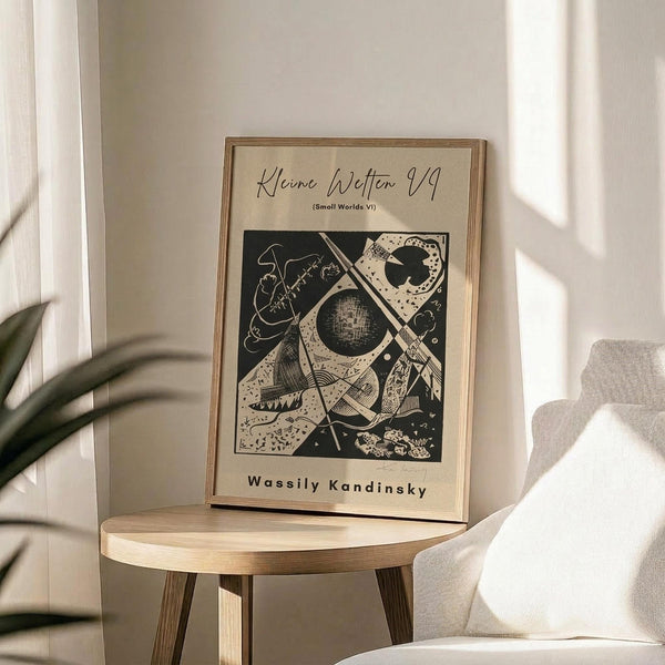 May include: Abstract geometric art print in light wood frame displaying black and white Kandinsky-style composition with circles, angular shapes, and organic forms. Framed poster sits on round wooden side table next to beige upholstered chair, with plant leaves visible in soft natural lighting against white walls.