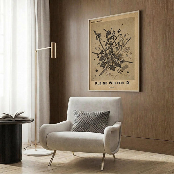 May include: Abstract geometric art print in warm beige and black tones displayed above a modern cream armchair with geometric patterned pillow. Vintage-style poster features intersecting lines, shapes, and patterns in Kandinsky's distinctive style, framed in light wood against wood-paneled wall. Golden floor lamp and dark side table with open book complete the contemporary reading nook setting with natural lighting from nearby window.