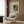 May include: Abstract geometric art poster by Wassily Kandinsky displayed in modern living room with warm wood paneling. Beige upholstered chair with geometric patterned pillow sits beside brass floor lamp and dark round side table with open book. Cream-colored poster features black and tan abstract shapes, circles, and lines in modernist style, mounted on wood wall near window with light curtains.